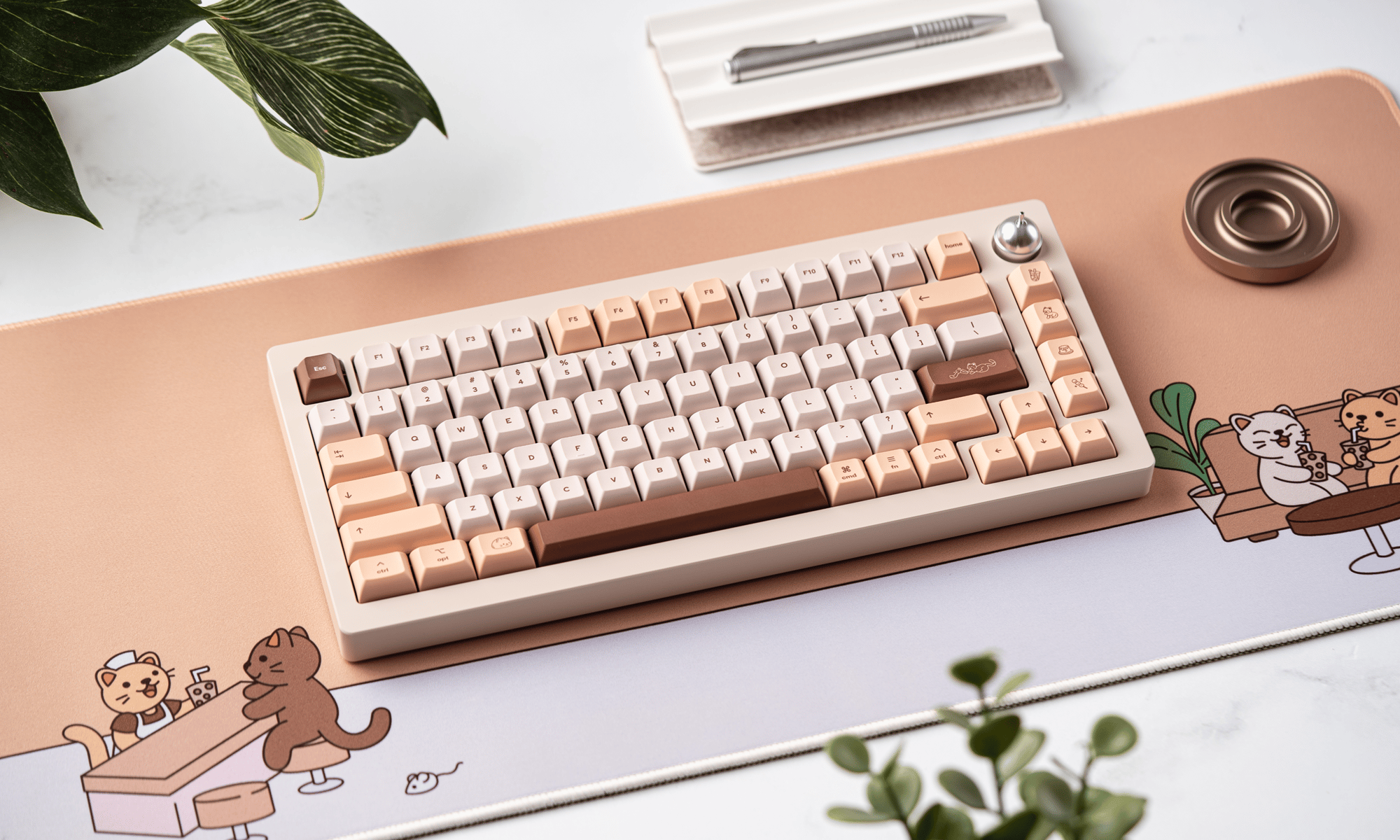 an angled birds-eye shot of Sprout 75 laying on the brown sugar boba deskpad. towards the upper edge of the image there's a notepad with a pen placed on it, to the side of the keyboard there's a gold screw tray, and just out of focus in the foreground there are a few plants.